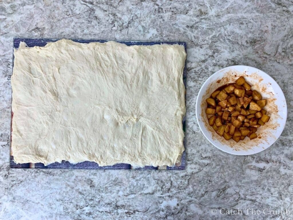 dough stretched out over a cutting board with apple mixture in a mixing bowl next to it