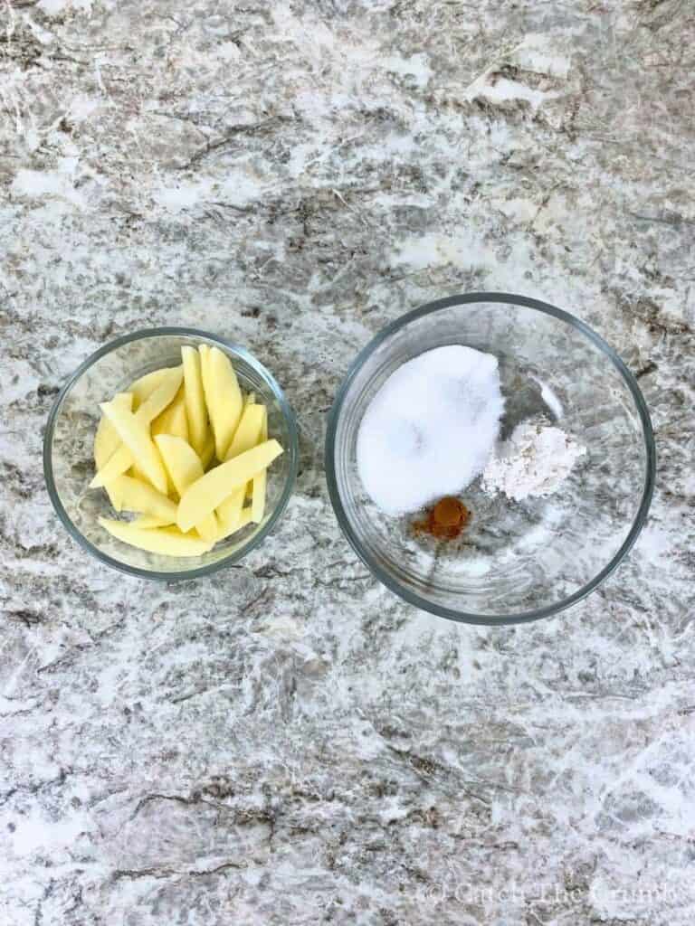 apple slices in a bowl next to a bowl containing granulated sugar, cinnamon, and flour
