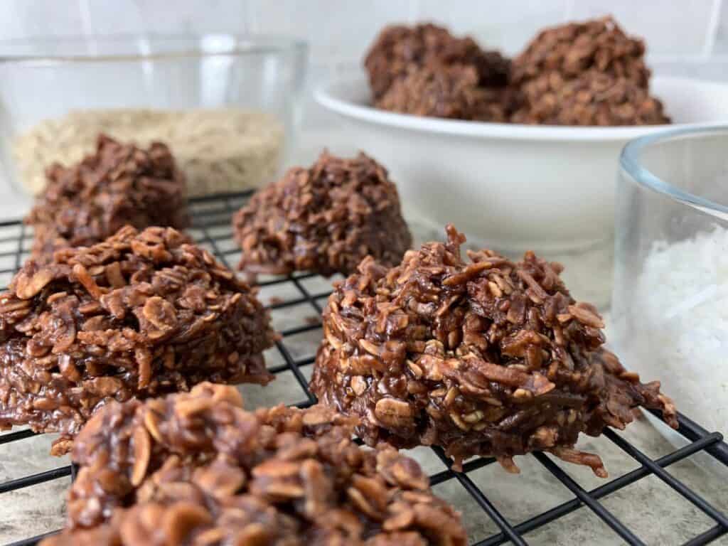 Close-up of chocolate coconut oat macaroons cooling on a wire rack, with bowls of oats and shredded coconut in the background.