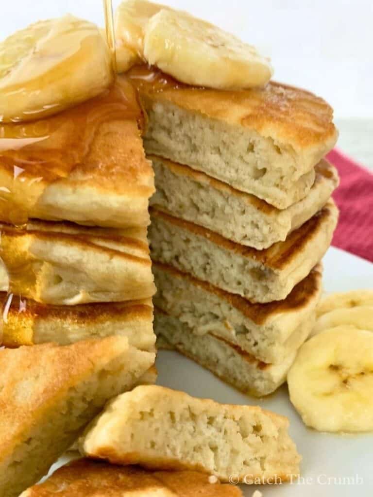 a stack of banana sourdough pancakes with pieces cut out and scattered on the plate in maple syrup