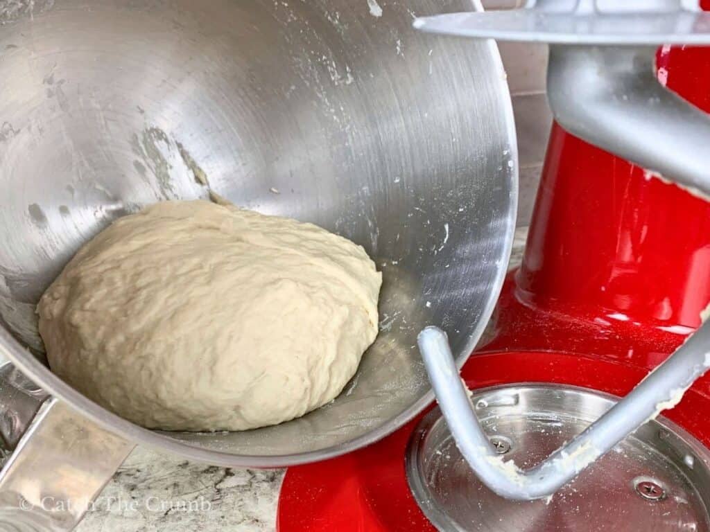 dough ball sitting in a mixing bowl next to a dough hook and mixer