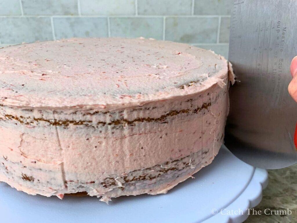 using a bench scraper to smooth the crumb coat layer of strawberry buttercream icing